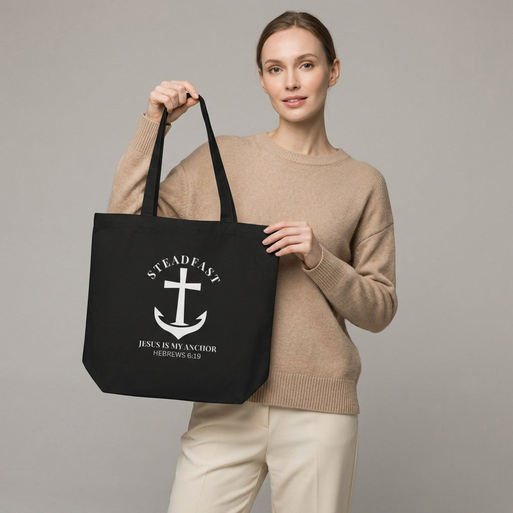 Model holding a black cotton tote bag printed with a white anchor cross and the words “STEADFAST” and “Jesus is my anchor — Hebrews 6:19.”