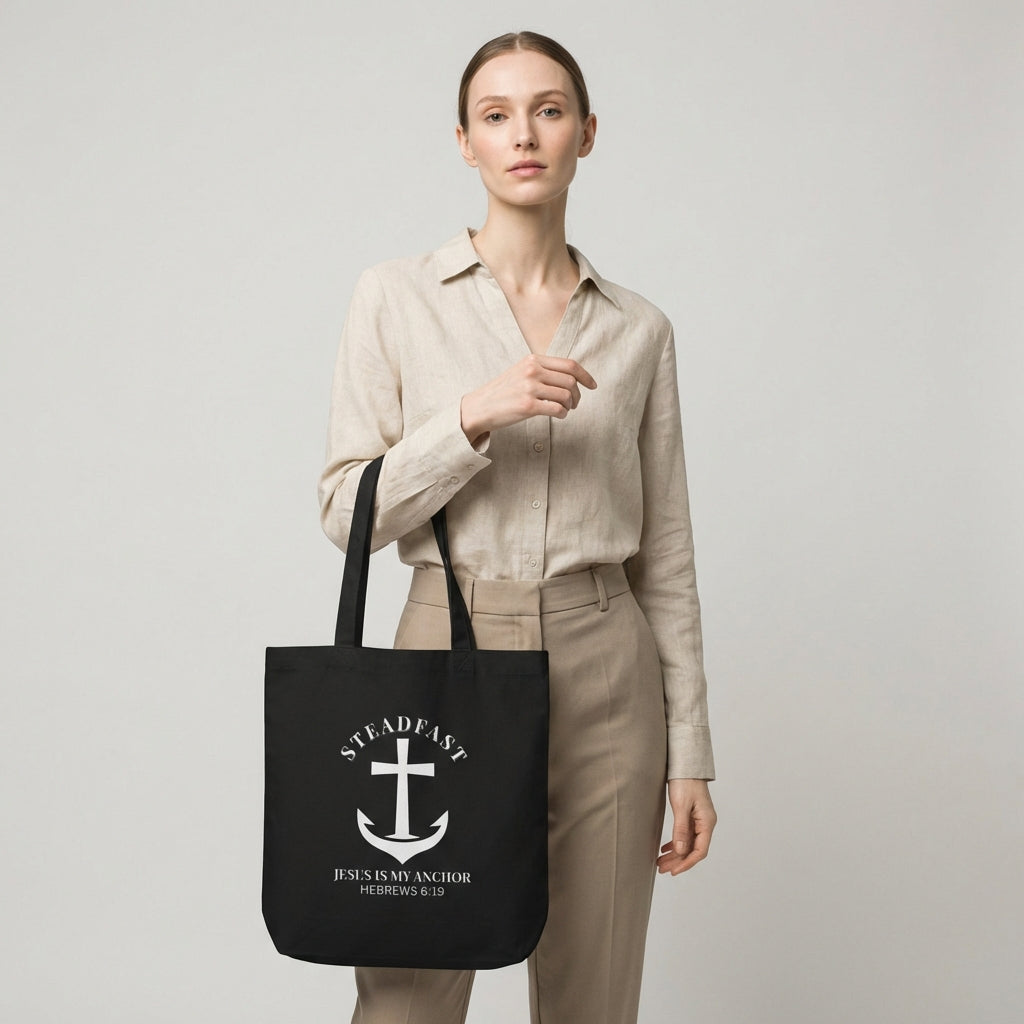 Model holding a black cotton tote bag printed with a white anchor cross and the words “STEADFAST” and “Jesus is my anchor — Hebrews 6:19.”