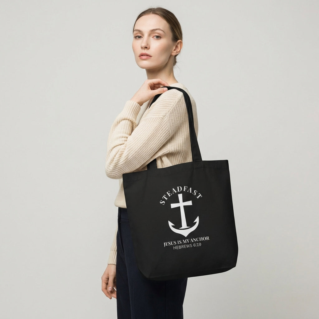Model holding a black cotton tote bag printed with a white anchor cross and the words “STEADFAST” and “Jesus is my anchor — Hebrews 6:19.”