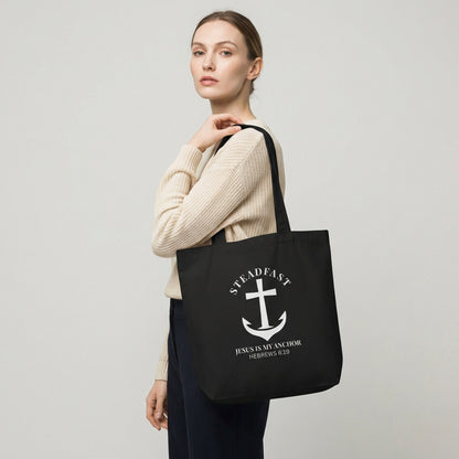 Model holding a black cotton tote bag printed with a white anchor cross and the words “STEADFAST” and “Jesus is my anchor — Hebrews 6:19.”