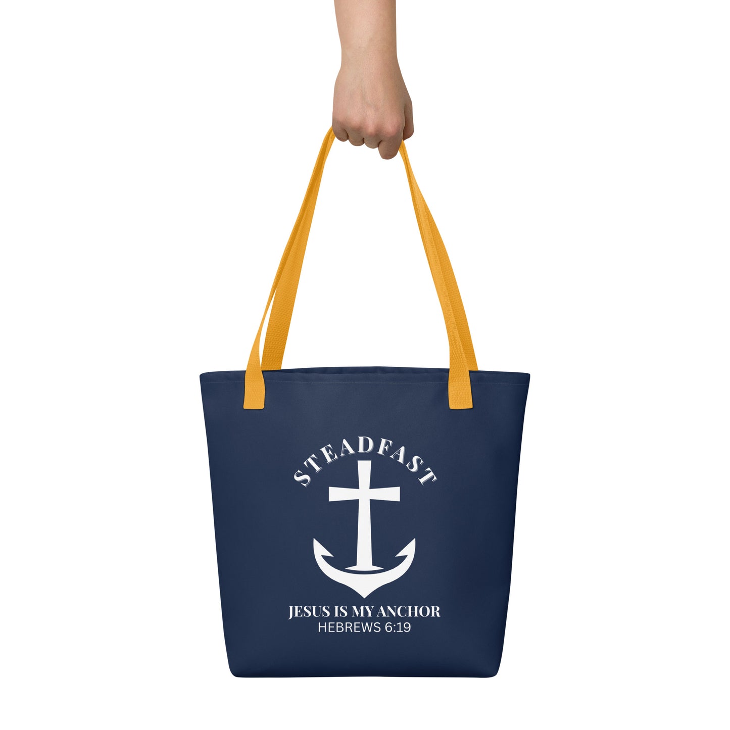 Tote bag held by hand with yellow straps, featuring a navy background and a white anchor cross graphic with the words “STEADFAST” and “JESUS IS MY ANCHOR” and the verse “HEBREWS 6:19.”