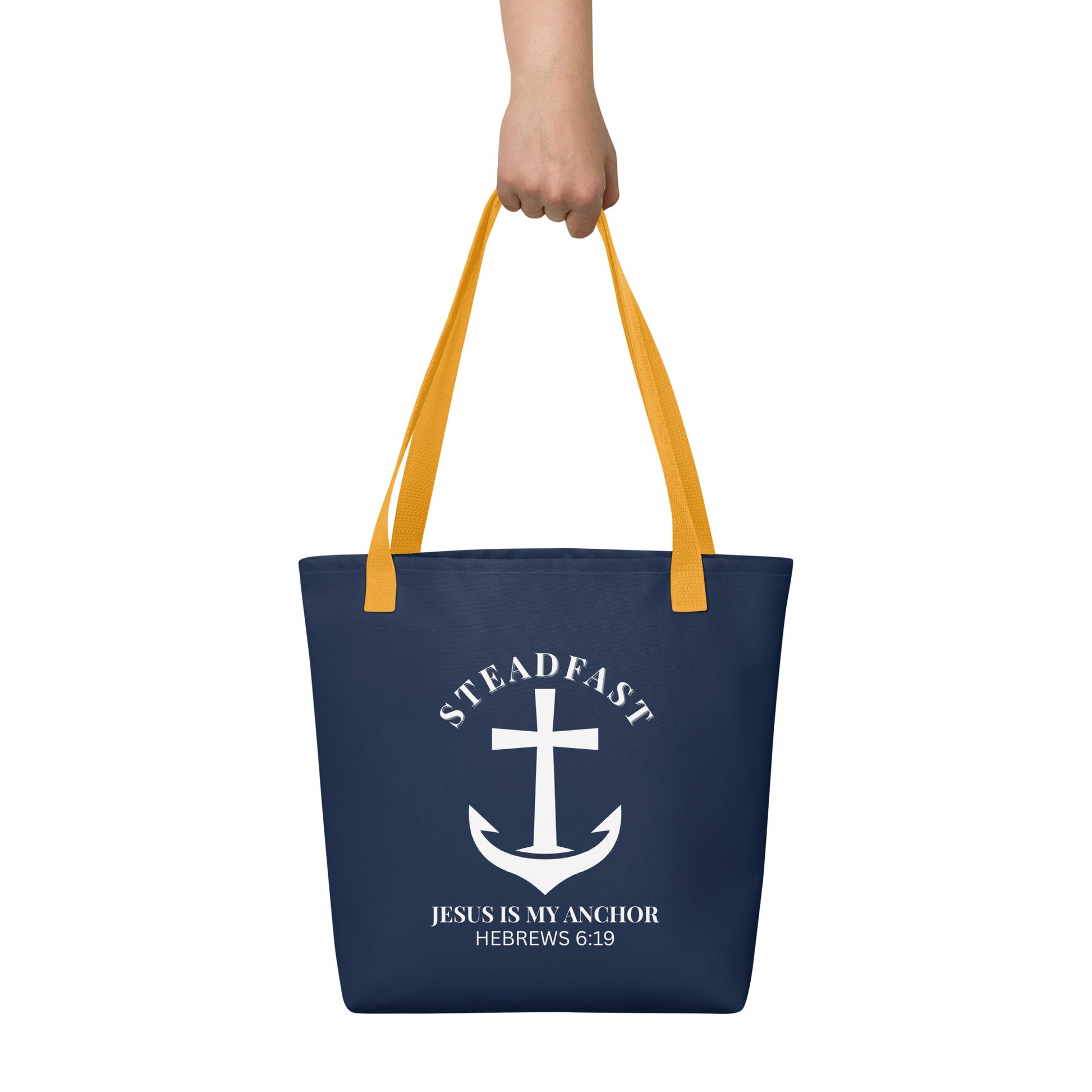 Tote bag held by hand with yellow straps, featuring a navy background and a white anchor cross graphic with the words “STEADFAST” and “JESUS IS MY ANCHOR” and the verse “HEBREWS 6:19.”