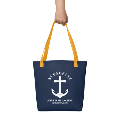 Tote bag held by hand with yellow straps, featuring a navy background and a white anchor cross graphic with the words “STEADFAST” and “JESUS IS MY ANCHOR” and the verse “HEBREWS 6:19.”