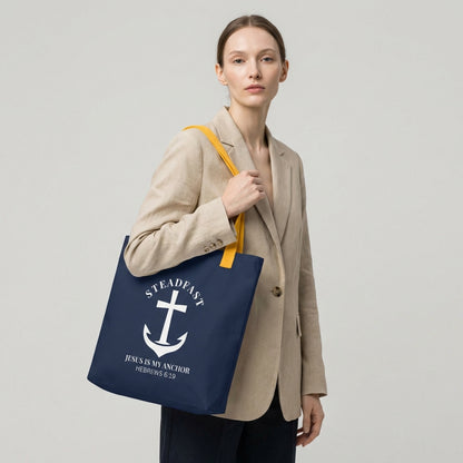 Model carrying a navy tote with mustard yellow straps, featuring a white anchor cross graphic and the message “STEADFAST — JESUS IS MY ANCHOR” (Hebrews 6:19) on the front.