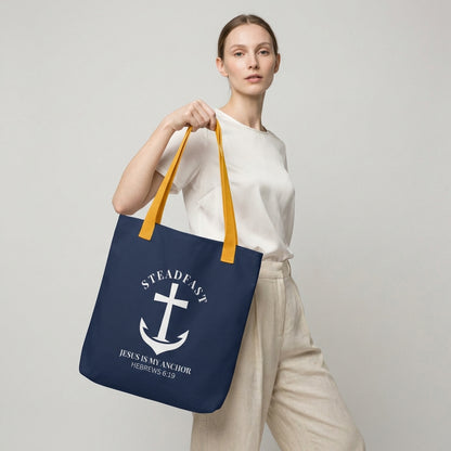 Model carrying a navy tote with mustard yellow straps, featuring a white anchor cross graphic and the message “STEADFAST — JESUS IS MY ANCHOR” (Hebrews 6:19) on the front.