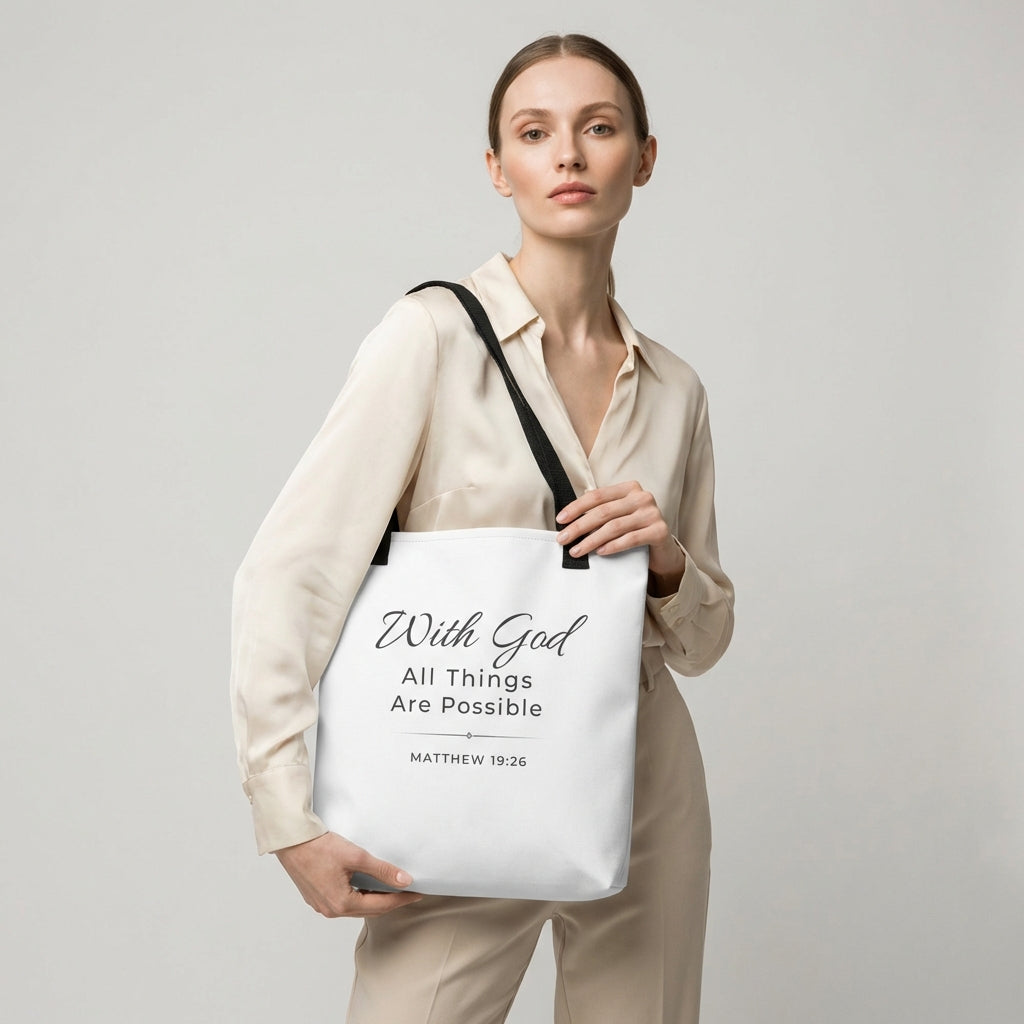 Lifestyle photo of a model holding a white tote bag with black handles featuring “With God all things are possible” and “Matthew 19:26” in dark gray.