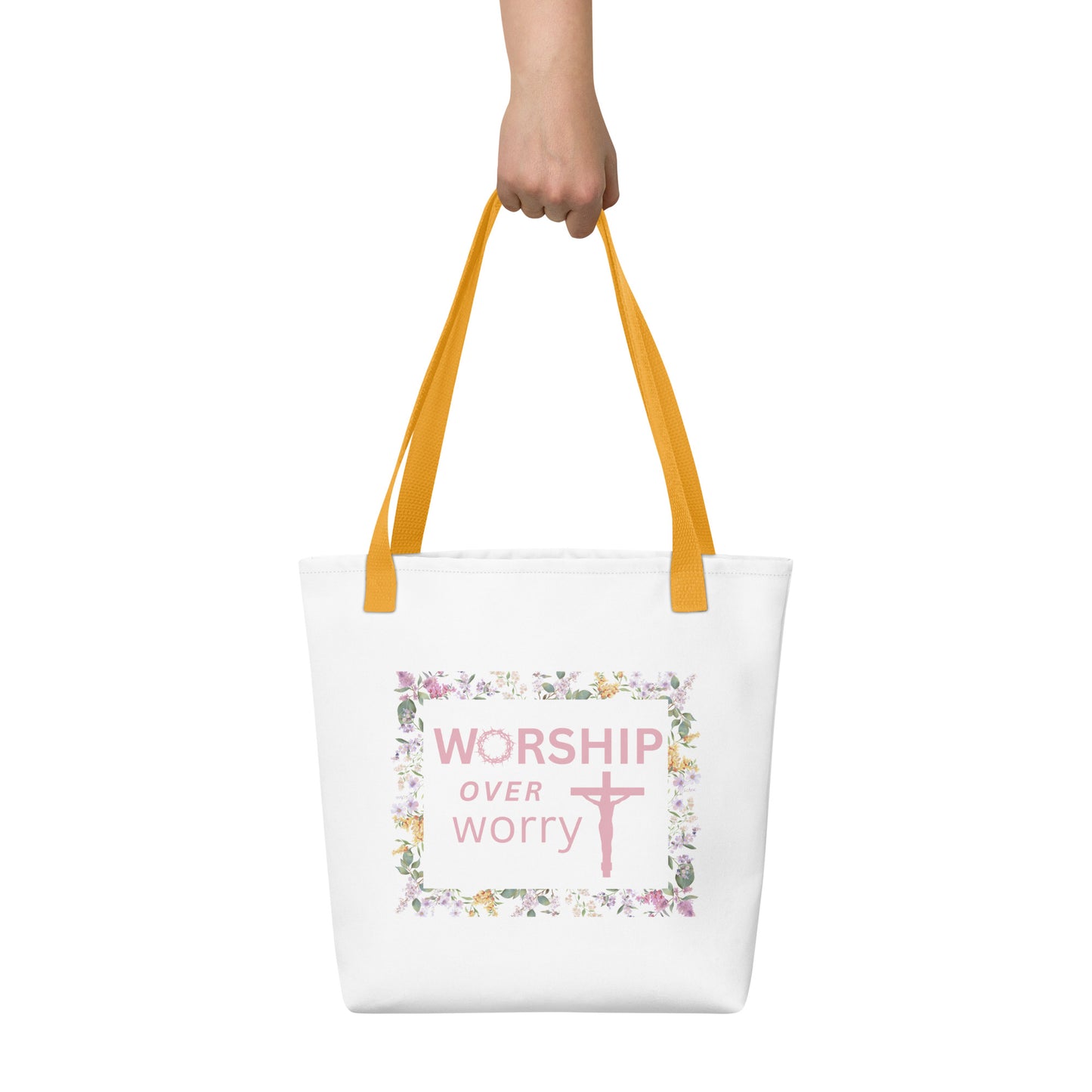Person holding a white tote with mustard-yellow handles, featuring the “WORSHIP over worry” message and floral border with cross.
