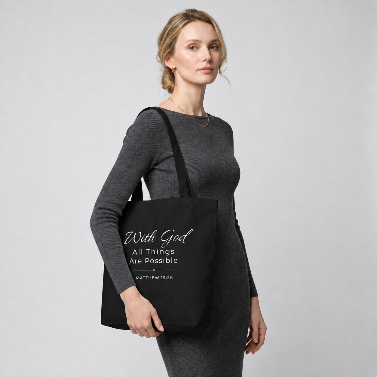 Model carrying a black cotton Christian tote bag with matching black handles and a white “With God All Things Are Possible” Matthew 19:26 design.