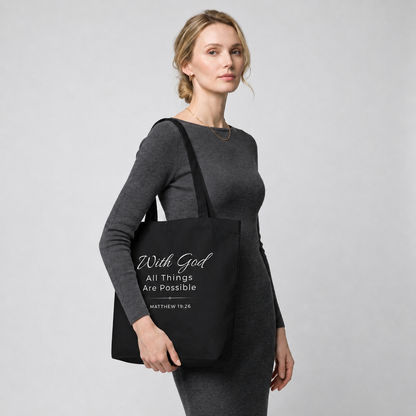 Model carrying a black cotton Christian tote bag with matching black handles and a white “With God All Things Are Possible” Matthew 19:26 design.